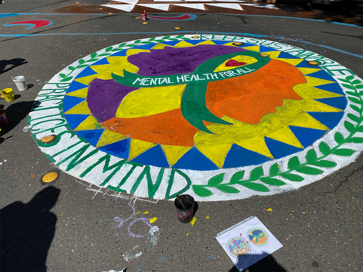 Colorful circular Earth Day street mural fully painted on pavement, featuring a stylized globe in bright purple, yellow, orange, and green with the words “Mental Health for All” across the center and “Keep Sustainability in Mind” around the edge.